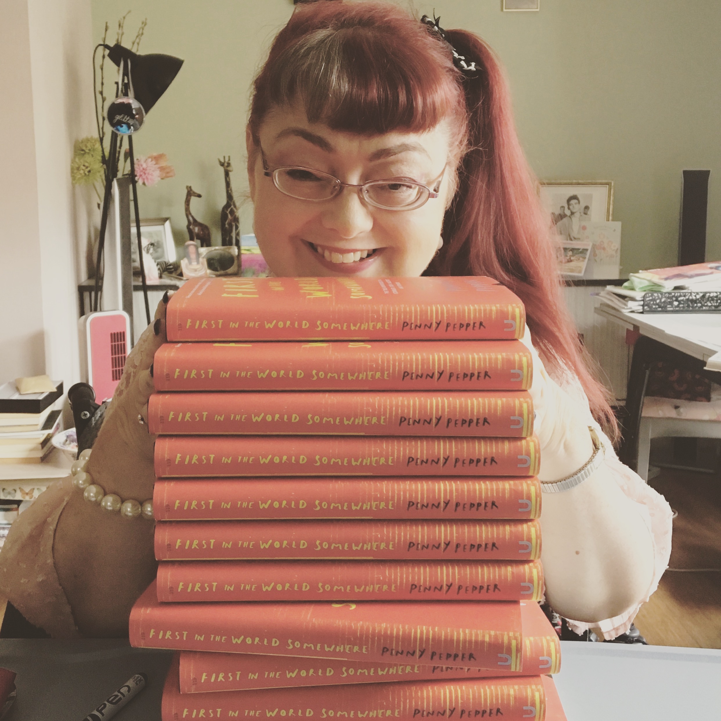 Penny is smiling and sits behind a pile of her memoir,  First in the World Somewhere. The  books are red with yellow writing. They are stacked  high with the spines showing to camera.