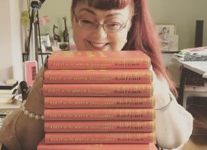 Penny is smiling and sits behind a pile of her memoir,  First in the World Somewhere. The  books are red with yellow writing. They are stacked  high with the spines showing to camera.