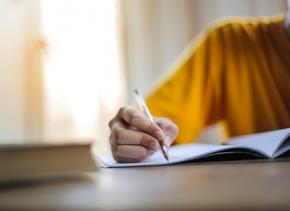 A person in a yellow jumper writes in a notebook lying on a desk.