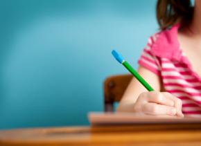 The image shows a child's hand holding a pencil and writing in a book.